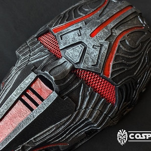 May include: A black and red detailed mask with a textured surface and a red mesh design. The mask has a futuristic and sci-fi aesthetic.