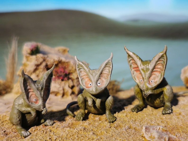 May include: Three small, green, and brown, fantasy creature figurines with large ears and wide eyes. The figurines are posed sitting on a sandy surface.