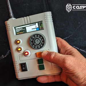 May include: A white and grey handheld device with a circular dial, a few buttons, and a switch. The device has a small screen with text that reads "SECURITY SYSTEM - 1234567890 - 0000000000 - NOSTALGIA 0".