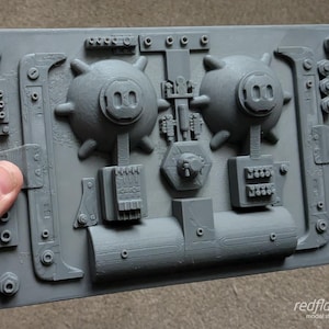 May include: A grey 3D printed wall panel with a steampunk design featuring two round, textured, bomb-like objects, a pipe, and various gears and levers.