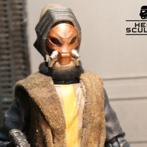 May include: A detailed action figure of a character with a copper-colored mask and head covering. The figure wears a brown and yellow outfit with a black collar. The text "HEAD SCULPTS" is visible.