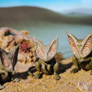 May include: Three whimsical, green and tan creature figurines with large, ear-like appendages and wide eyes. They are posed on a sandy surface with a blurred background of water and a rocky outcropping. The creatures have a unique, otherworldly appearance.
