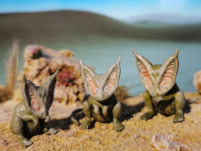 May include: Three whimsical, green and tan creature figurines with large, ear-like appendages and wide eyes. They are posed on a sandy surface with a blurred background of water and a rocky outcropping. The creatures have a unique, otherworldly appearance.