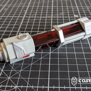 Puede incluir: Un accesorio cilíndrico con un núcleo rojo oscuro translúcido encerrado en un marco blanco mate con detalles metálicos plateados. El objeto tiene un detalle circular en la parte superior y una estructura de base rectangular con líneas limpias.