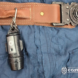 Può includere: Un accessorio cilindrico in metallo nero con un fermaglio argentato e un simbolo bianco sul lato. L'accessorio è attaccato a una cintura in pelle marrone con una fibbia in metallo.