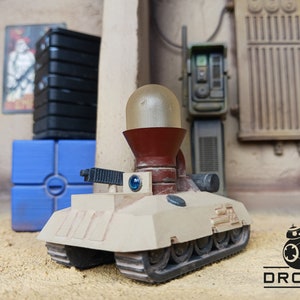 May include: A beige toy tank with a red and clear dome on top. The tank has tracks and a small gun on the front. The tank is sitting on a sandy surface. The word "DROIDS" is printed in black on a white background in the lower right corner of the image.
