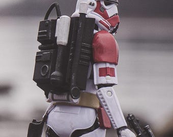 Puede incluir: Una figura de acción de stormtrooper blanca y roja que lleva una mochila negra con un accesorio cilíndrico. La figura presenta marcas rojas en el casco y la hombrera, un cinturón utilitario color canela y sostiene un bláster negro.