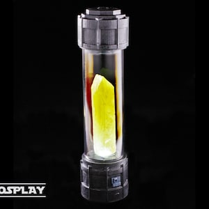 Puede incluir: Un tubo de vidrio transparente con un cristal amarillo en su interior. El tubo está encerrado en un marco de metal negro con un interruptor en el lateral. El cristal brilla con una luz amarilla.
