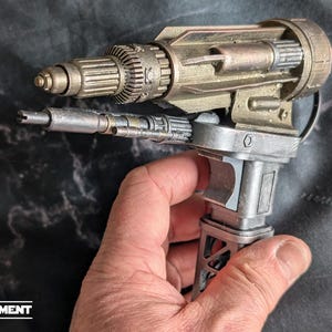 May include: A metallic, futuristic-looking prop gun with intricate details. The gun features a cylindrical barrel, a ribbed grip, and various mechanical components. The color scheme is primarily silver and gold, with a weathered appearance. The word "EQUIPMENT" is at the bottom.