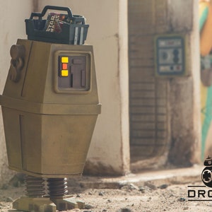 Può includere: Un piccolo robot marrone con una cassetta degli attrezzi in cima. Il robot ha una luce gialla e rossa sul davanti. Il robot è in piedi su una superficie sabbiosa.
