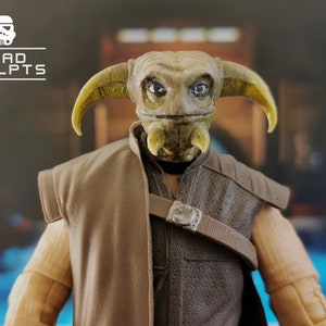 May include: A green and brown action figure with a large head and pointed ears. The figure is wearing a brown vest and a brown belt with a silver buckle. The figure is from the Star Wars franchise.