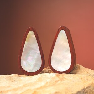 May include: A pair of teardrop earrings. Each earring has a dark reddish-brown wooden frame with a pearlescent, iridescent inlay. The earrings are presented on a light brown, textured surface against a gradient brown background.