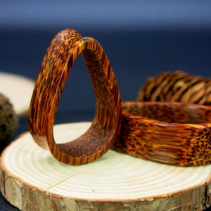 May include: Two wooden rings with a natural wood grain pattern. The rings are brown and orange with a reddish hue. The rings are on a wooden surface.