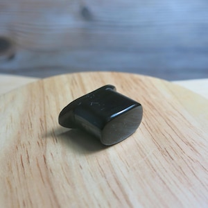 May include: A small, black, polished plug, possibly for ear piercing, sits on a light-coloured wooden surface. The plug has a rounded, rectangular shape with a slight curve and a small hole at the top.