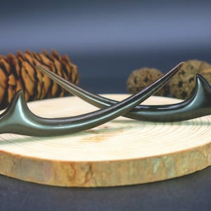 May include: Two dark brown hair sticks with a curved design, resting on a wooden slice. A pine cone and other natural elements are in the background. The hair accessories have a smooth, polished finish.