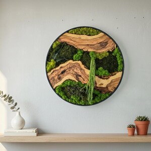 May include: Round wall art with a black frame, featuring a natural moss and wood design. The artwork incorporates various shades of green moss and light brown wood elements. A decorative piece for home decor.