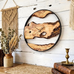 May include: A circular wall clock featuring a natural wood face and a black metal frame. The clock face is crafted from light brown, textured wood with dark brown bark edges. Black clock hands indicate the time. It's mounted on a white wooden wall.