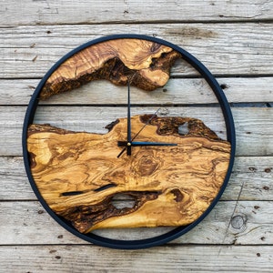 Op de afbeelding: Een ronde houten klok met een zwart frame. De wijzerplaat van de klok is gemaakt van een natuurlijke houtschijf met een uniek nerfpatroon. De wijzers van de klok zijn zwart en de wijzerplaat van de klok heeft een gouden accent.