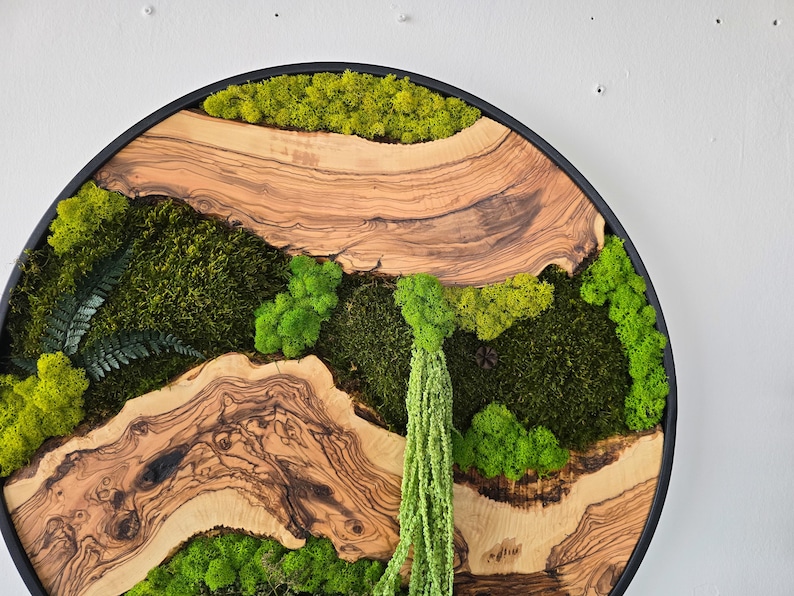 May include: Circular wall art featuring a natural landscape design. The artwork combines light brown wood with green moss and a black frame. The design includes a variety of textures and shades of green.