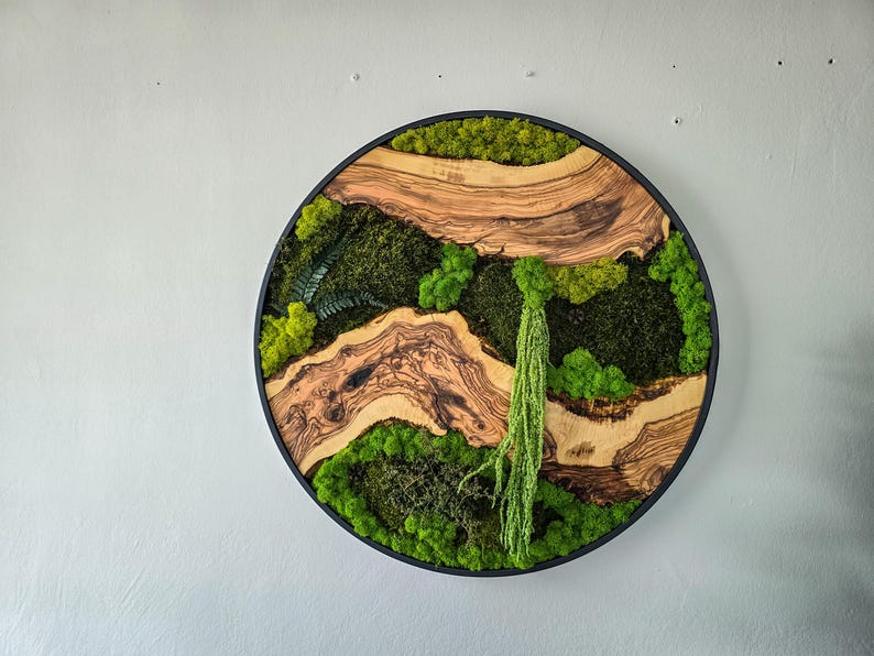 May include: Round wall art featuring a natural landscape design. The artwork combines wood and moss, with a black frame. The wood has a light brown color with dark brown grain patterns. The moss is various shades of green.