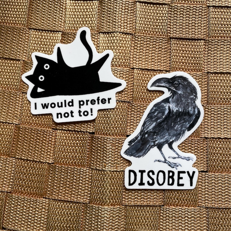 May include: Two stickers on a textured surface. One sticker features a black cat with the text "I would prefer not to!". The other sticker is a detailed illustration of a black crow with the word "DISOBEY".