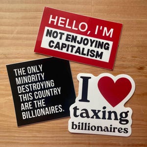 May include: Three stickers with bold text on a wooden surface. One red sticker reads "HELLO, I'M NOT ENJOYING CAPITALISM." A black sticker says "THE ONLY MINORITY DESTROYING THIS COUNTRY ARE THE BILLIONAIRES." A white sticker has a red heart and says "I ❤️ taxing billionaires."