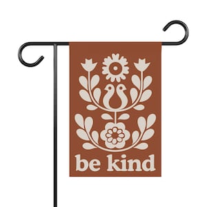 May include: A garden flag with a rust-colored background and a cream-colored floral design. The design includes flowers, leaves, and two bird-like figures. The words "be kind" are printed at the bottom in cream-colored letters. The flag is attached to a black metal stand.