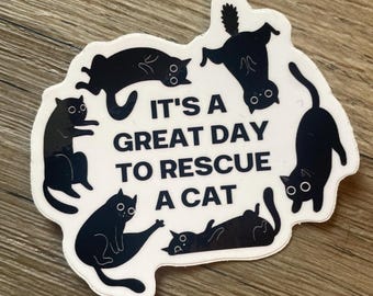 May include: A white sticker with the text "IT'S A GREAT DAY TO RESCUE A CAT" surrounded by black cat illustrations in various poses. The sticker is on a wooden surface.
