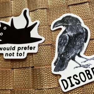 May include: Two stickers on a textured surface. One features a black cat with the text "I would prefer not to!". The other is a detailed illustration of a raven with the word "DISOBEY".