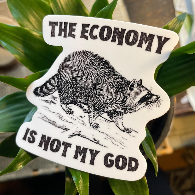 May include: A white sticker with a black and white illustration of a raccoon walking on a log. The sticker has the text "The Economy is Not My God" in black.