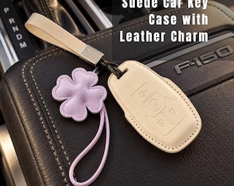 May include: A beige suede car key case featuring a matching beige wrist strap and a light purple four-leaf clover leather charm. The case has embossed button icons and is attached to a metal connector. Text reads: Suede Car Key Case with Leather Charm.