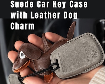 May include: A grey suede car key case with a Volkswagen logo embossed on the surface, attached to a brown leather dog charm by a black cord. The text "Suede Car Key Case with Leather Dog Charm" is displayed at the top of the image.
