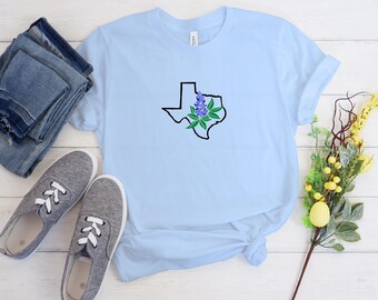 May include: A light blue short-sleeve t-shirt featuring a black outline of the state of Texas with a purple bluebonnet flower and green leaves centered on the chest. The shirt is displayed alongside a pair of grey sneakers and folded denim jeans.