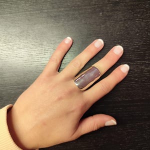 Könnte beinhalten: Ein silberfarbener Ring mit einem rechteckigen, irisierenden lila Stein. Der Ring wird an einer Hand mit manikürten Nägeln getragen. Das Design ist schlicht und modern, für den Alltag geeignet.