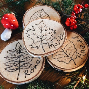 May include: Four wooden coasters with different leaf designs burned into the surface. The coasters are labelled "Maple", "Rowan", "Hazel", and "Maple".