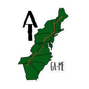 May include: A green outline of the northeastern United States with a red line tracing the Appalachian Trail. The text "GA-ME" is below the map. An arrow pointing upwards is in the top left corner.