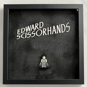 Może przedstawiać: Czarna, oprawiona ramą, ramka z cieniem z białym tłem. Tytuł "EDWARD SCISSORHANDS" jest napisany białymi literami. Figurka Lego Edwarda Scissorhands znajduje się w środku ramki.