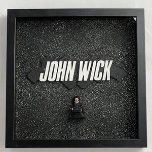 May include: A black framed shadow box with a black and white speckled background. The words "JOHN WICK" are printed in white on a black background. A black Lego figure of John Wick is in the center of the box.