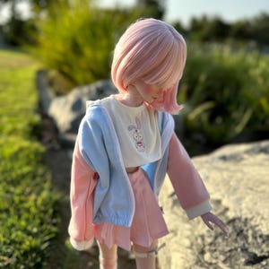 Könnte beinhalten: Eine Puppe mit rosa Haaren und einem passenden Outfit. Die Puppe trägt ein weißes Hemd mit einem Hasenmuster, eine hellblaue und rosa Jacke und einen rosa Rock. Die Puppe steht draußen an einem sonnigen Tag.
