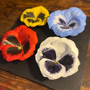 May include: Four colorful pansy flowers arranged on a black surface. The flowers are in shades of red, yellow, blue, and white, each with unique dark markings in the center. The petals have a delicate, textured appearance.