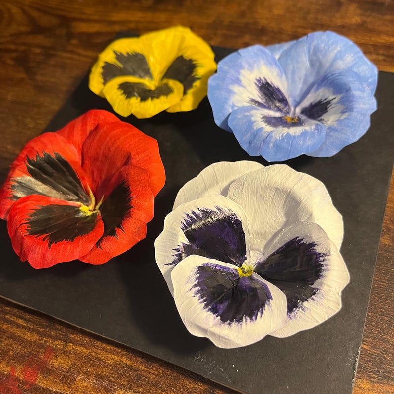 May include: Four colorful pansy flowers arranged on a black surface. The flowers are in shades of red, yellow, blue, and white, each with unique dark markings in the center. The petals have a delicate, textured appearance.