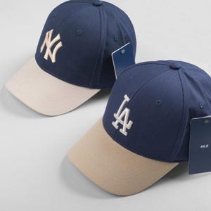 Könnte beinhalten: Zwei Baseballkappen mit einer dunkelblauen Krone und einem beigefarbenen Schirm. Die Kappe links hat ein weißes "NY"-Logo. Die Kappe rechts hat ein weißes "LA"-Logo.