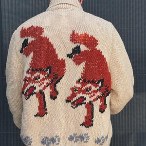 May include: A beige knit cardigan featuring a shawl collar and a back design of two identical rust-colored, stylized animal figures with black outlines and red accents. The bottom hem displays a repeating pattern of small gray shapes.