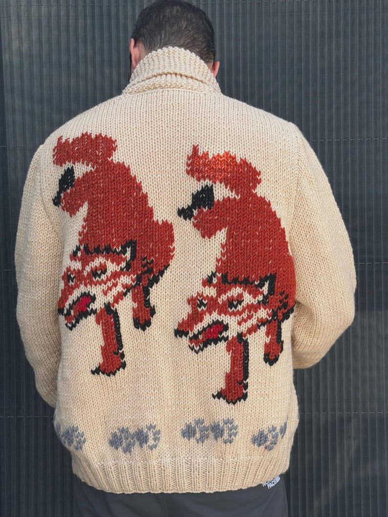 May include: A beige knit cardigan featuring a shawl collar and a back design of two identical rust-colored, stylized animal figures with black outlines and red accents. The bottom hem displays a repeating pattern of small gray shapes.