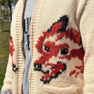 May include: A cream-colored knit zip-up cardigan featuring a prominent red and black fox graphic on the front panel. The garment includes a ribbed collar, a full-length zipper, and a patch pocket. A grey t-shirt with partial black text is visible underneath.