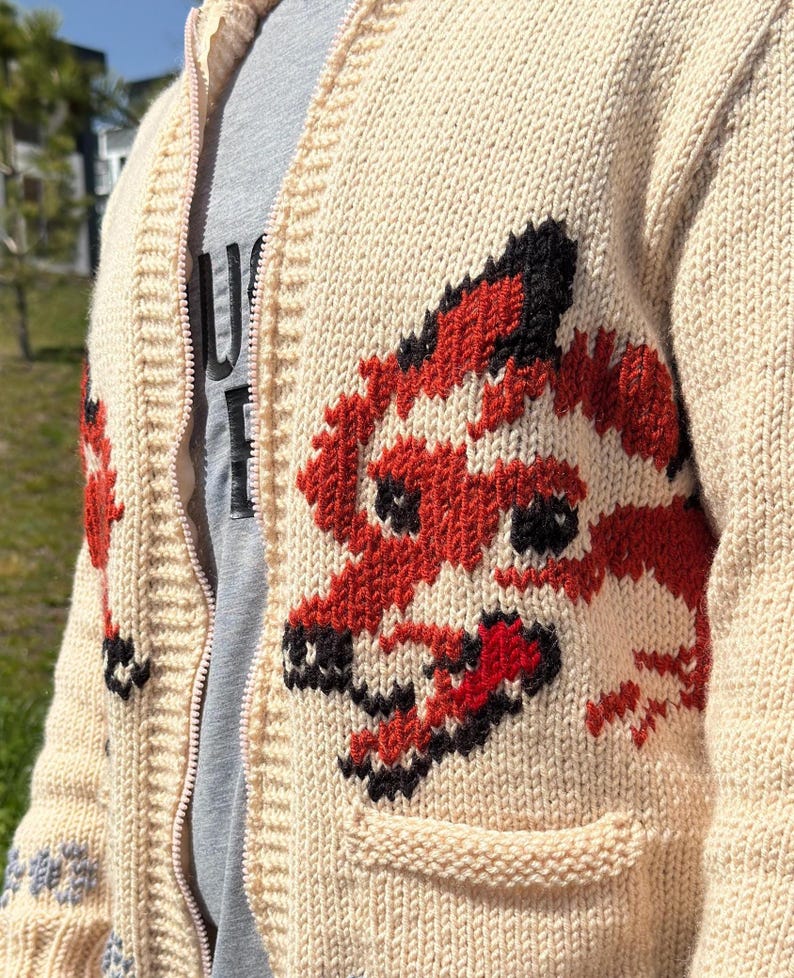 May include: A cream-colored knit zip-up cardigan featuring a prominent red and black fox graphic on the front panel. The garment includes a ribbed collar, a full-length zipper, and a patch pocket. A grey t-shirt with partial black text is visible underneath.