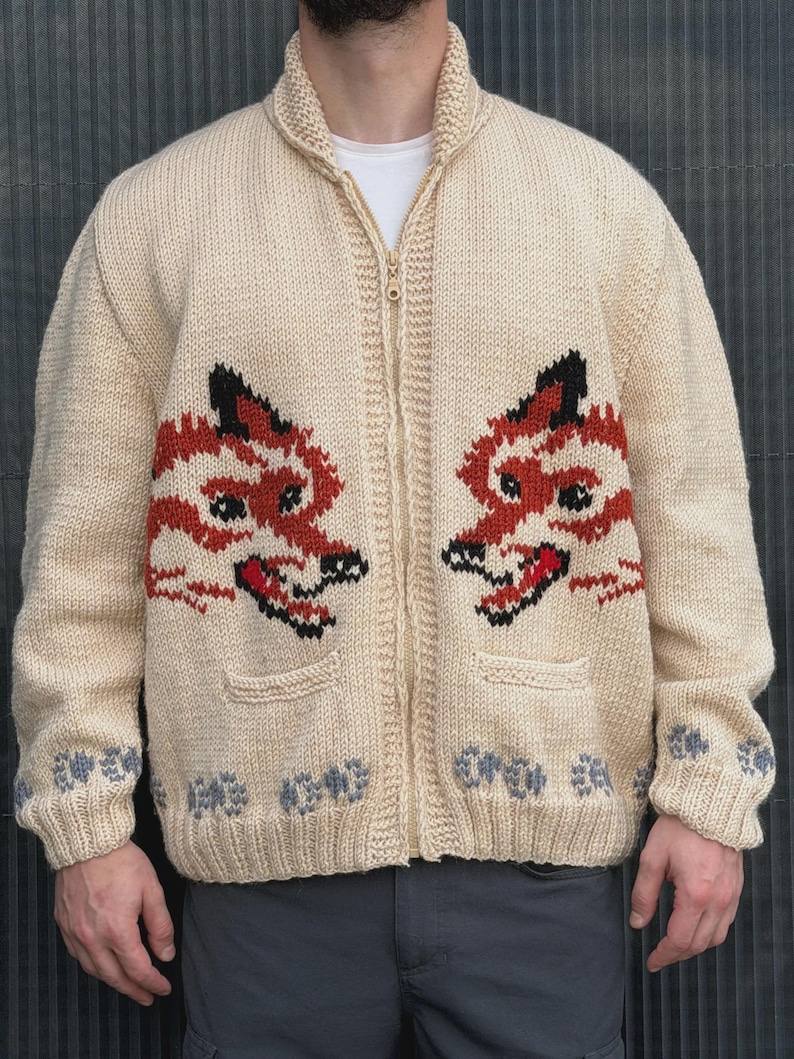 May include: A cream-colored knit zip-up cardigan featuring two large fox head motifs in orange, black, and white on the front panels. The sweater includes a high collar, a central zipper closure, two front pockets, and a grey paw print pattern at the hem.