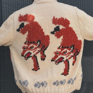 May include: A beige knit cardigan featuring a shawl collar and a back design of two stylized red and black foxes. The hem is decorated with a row of small grey paw prints, and the garment includes a small black logo patch on the lower right side.