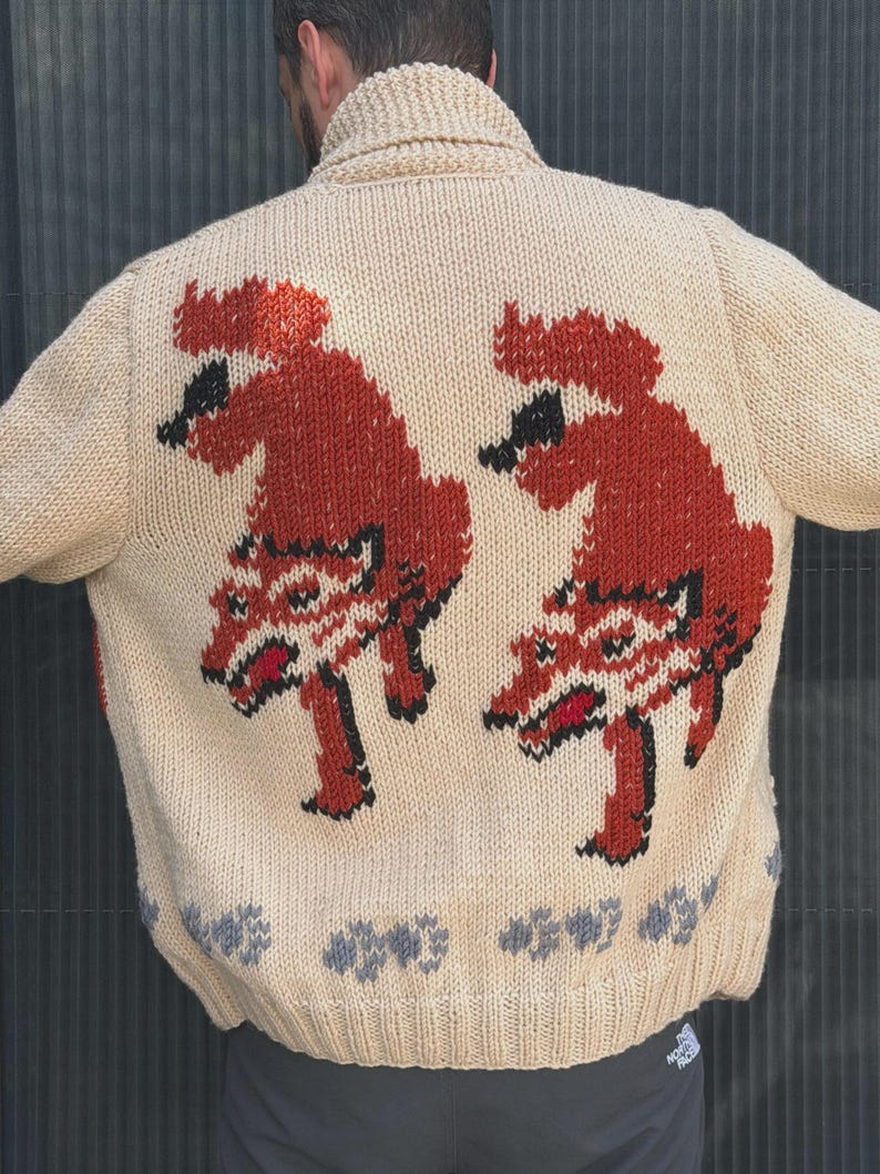 May include: A beige knit cardigan featuring a shawl collar and a back design of two stylized red and black foxes. The hem is decorated with a row of small grey paw prints, and the garment includes a small black logo patch on the lower right side.