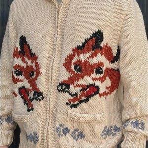 May include: A beige knit zip-up sweater featuring two large fox motifs on the front panels. The foxes are rendered in orange, black, and white yarn. The sweater includes two front pockets and a row of small grey paw print patterns along the hem.